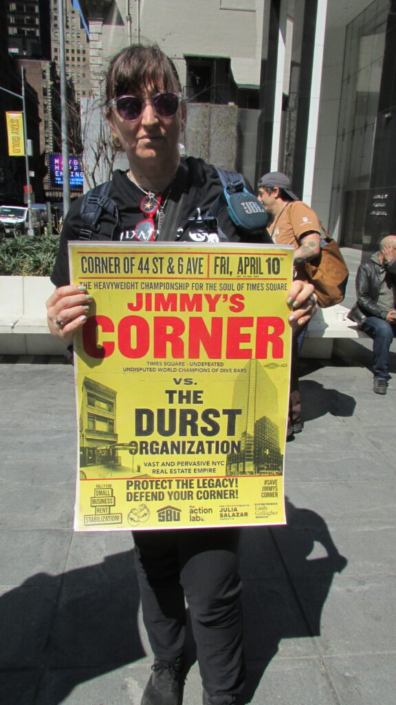 Person with sunglasses holds a large yellow poster for Jimmy's Corner in a city street.